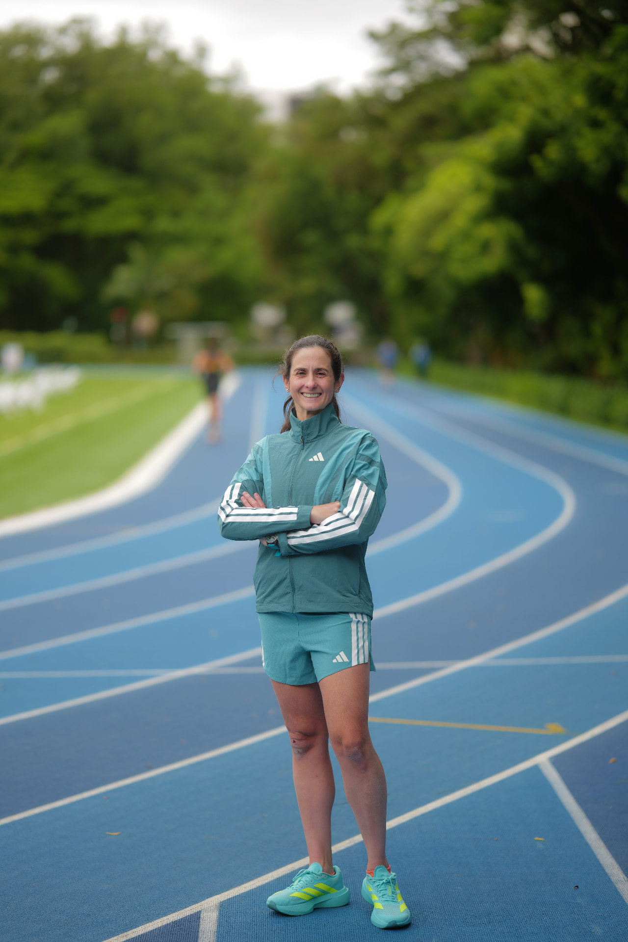 Ana Paula de corpo inteiro, de braços cruzados, em uma pista de atletismo azul