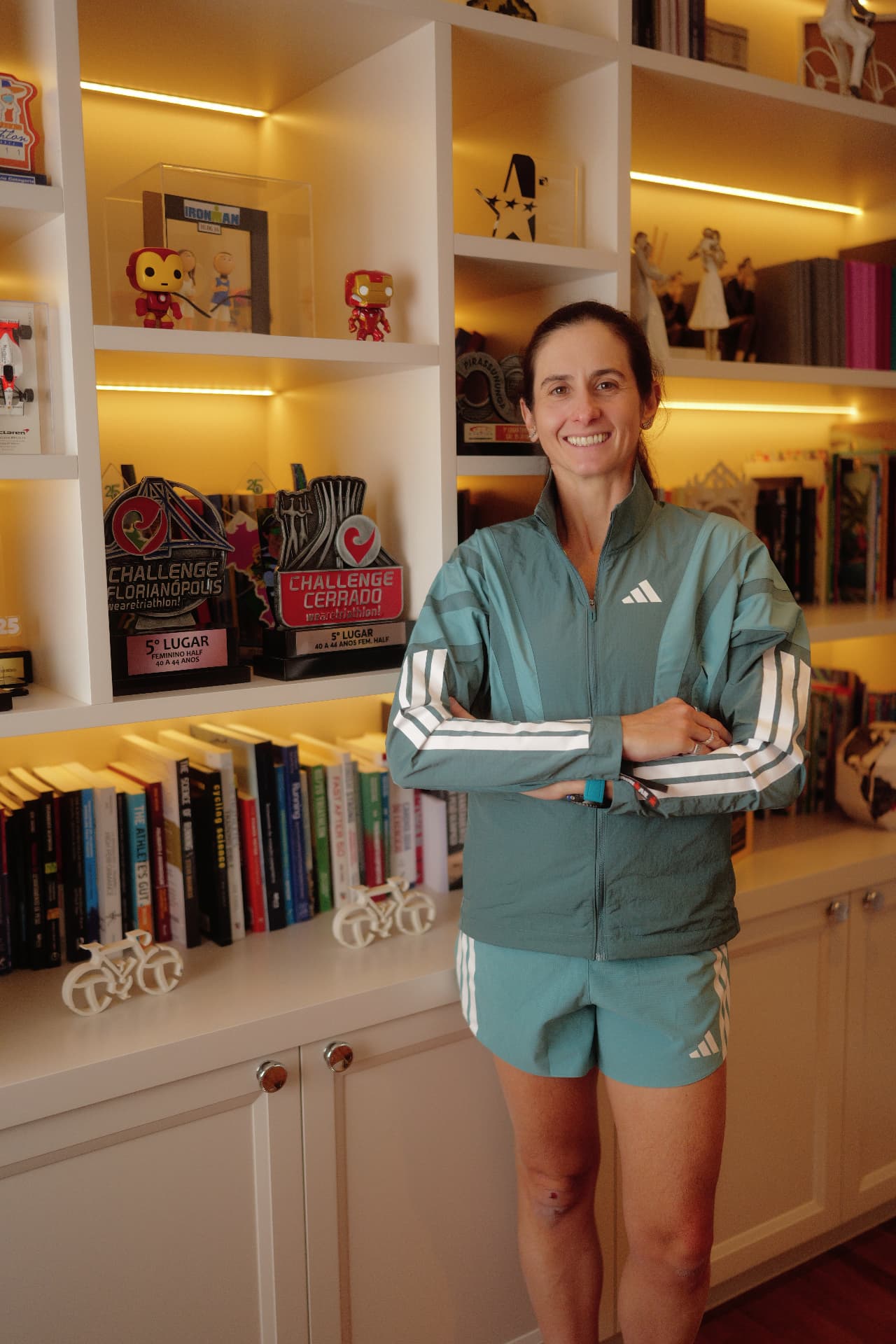 Ana Paula de braços cruzados, sorrindo, em frente a uma estante com troféus de triathlon e livros sobre corrida e ciclismo